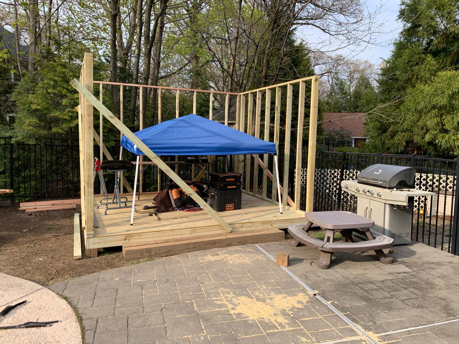 A framed shed floor and walls on a patio, with a blue canopy, a picnic table and a grill